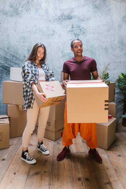 A professional removal worker standing inside a home during a house relocation process, next to several filled cardboard boxes of various sizes, some sealed with red packing tape. The worker, dressed 