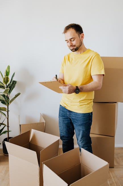 A man with short dark hair and a beard, dressed in a yellow t-shirt and blue jeans, is standing indoors next to several open cardboard moving boxes. He is holding a clipboard and a pen, appearing to c